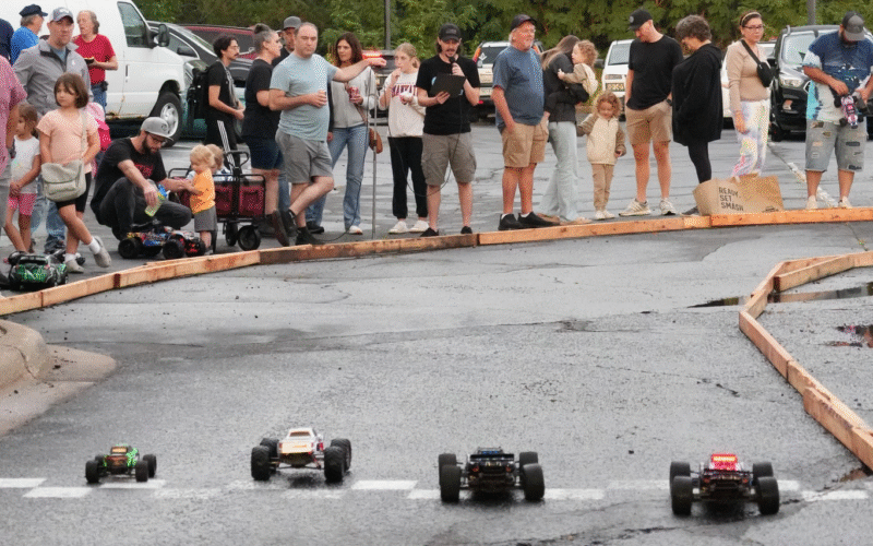 four radio controlled cars lined up on a race track in a parking lot full of adults and kids
